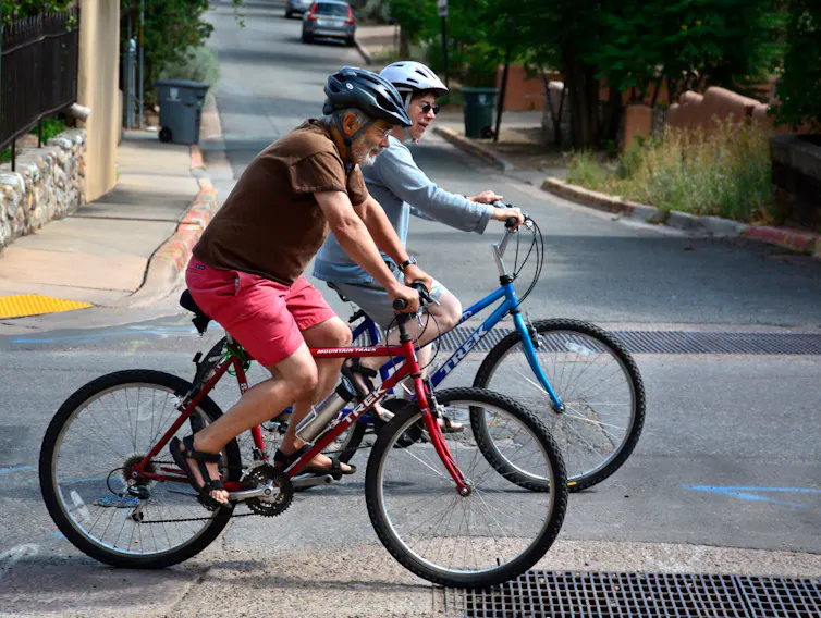 An elderly man and an elderly woman ride their bikes on a street while wearing helmets.