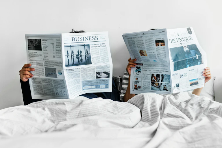 Two people in bed reading different sections of a newspaper.