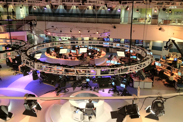 A room with large screens, computers and several tables showing a newsroom setup.