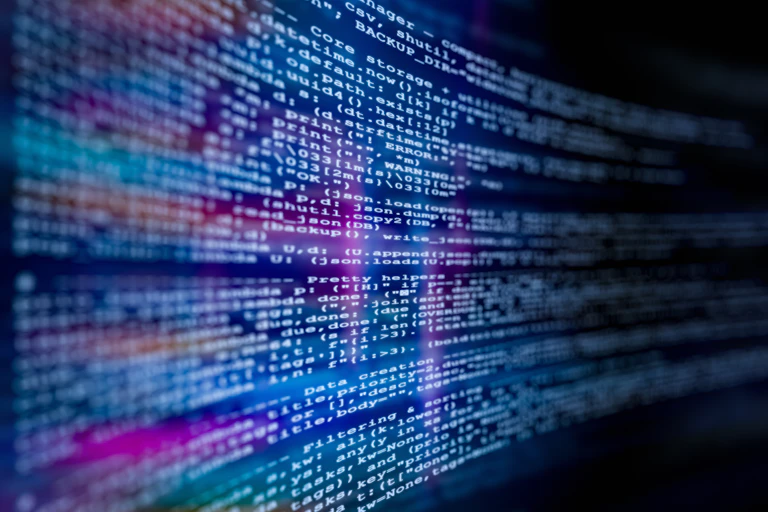 Close-up view of computer code displayed on a digital screen, with blurred lines of text and glowing blue and purple light reflections.