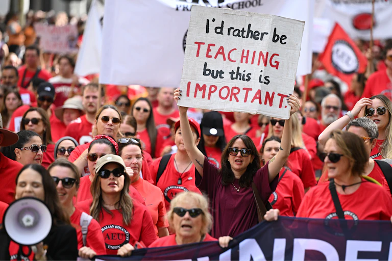 A woman in a large crowd, holding up a sign saying I'd rather be teaching but this is important