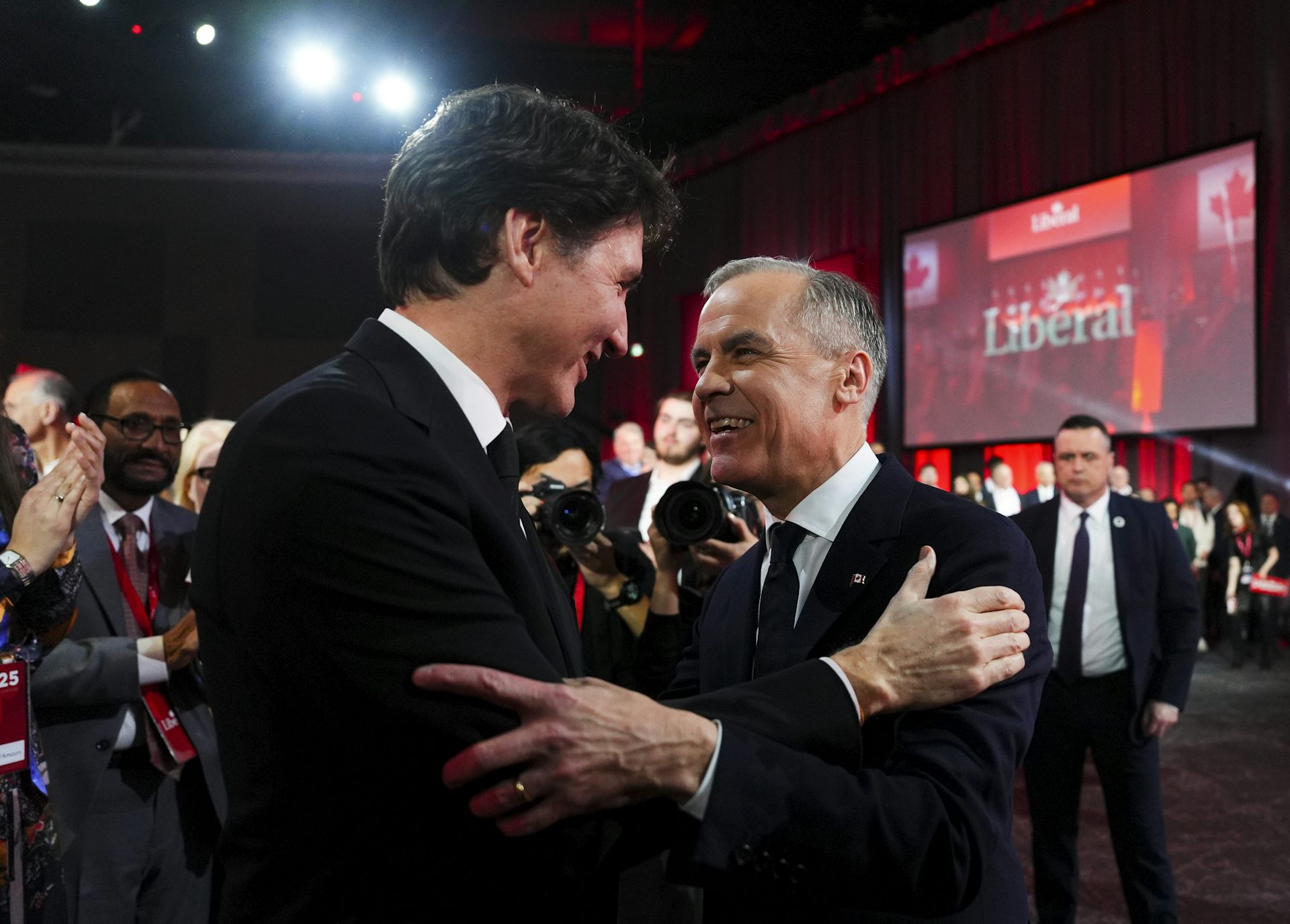 Dos hombres, uno más joven y alto con cabello oscuro, el otro con cabello corto y gris, se abrazan.