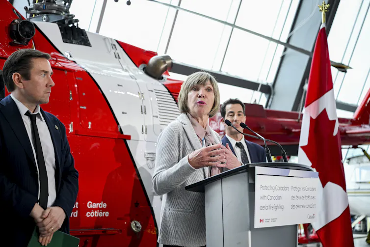 una mujer con traje blanco habla en un podio frente a un helicóptero y una bandera canadiense en un poste
