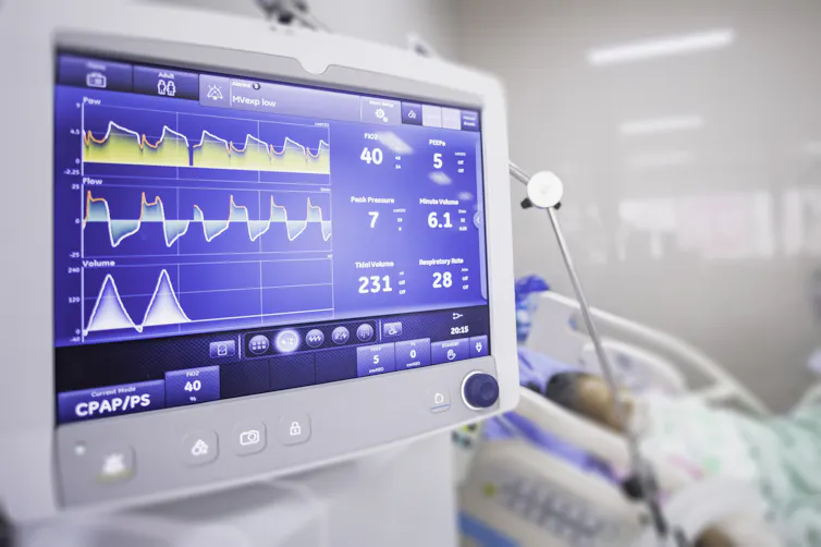 A ventilator connected to a patient shows vital readings on a blue screen in a hospital room.
