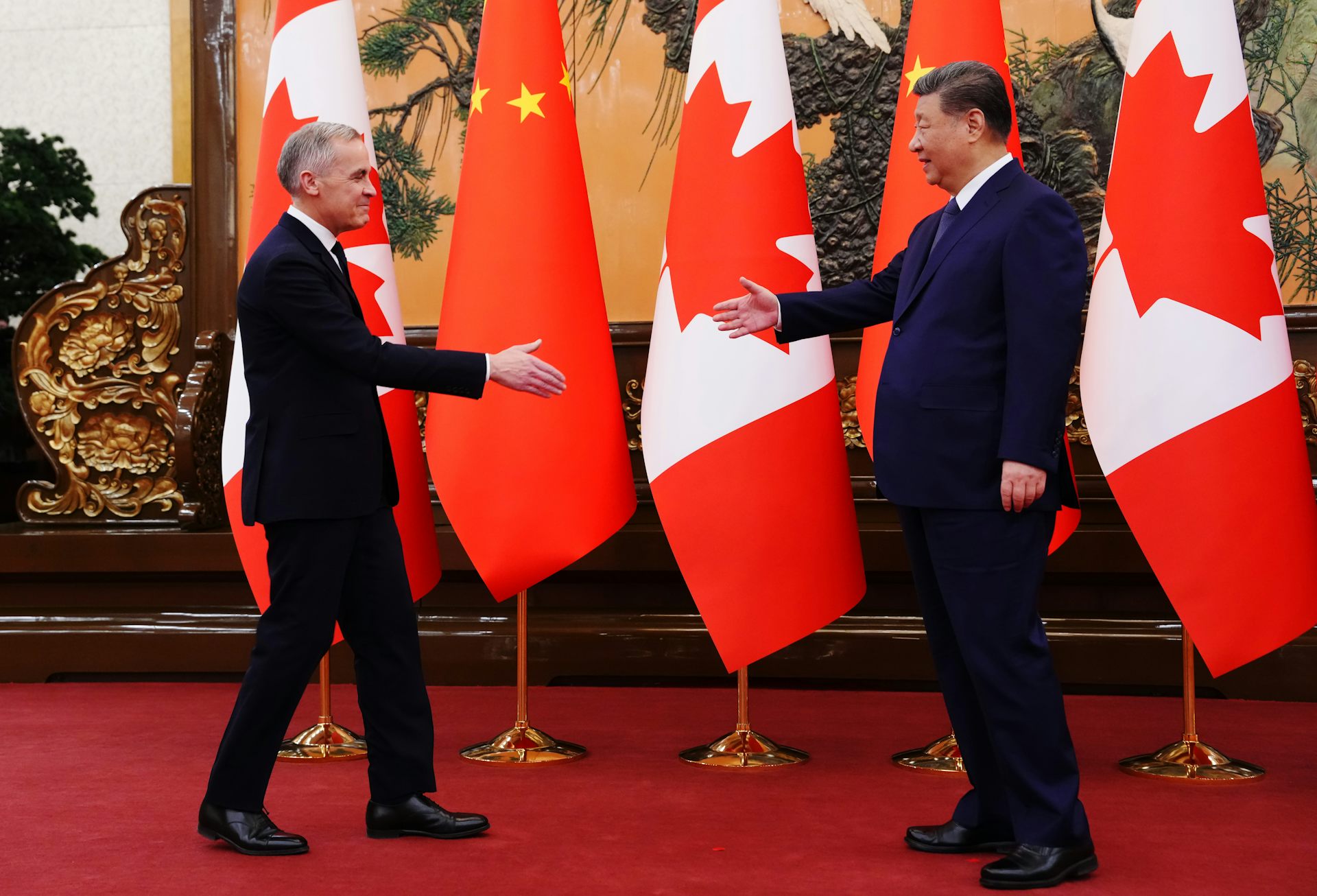 Dos hombres de mediana edad, uno blanco y otro chino, se acercan para darse la mano mientras están parados frente a una fila de banderas canadienses y chinas.