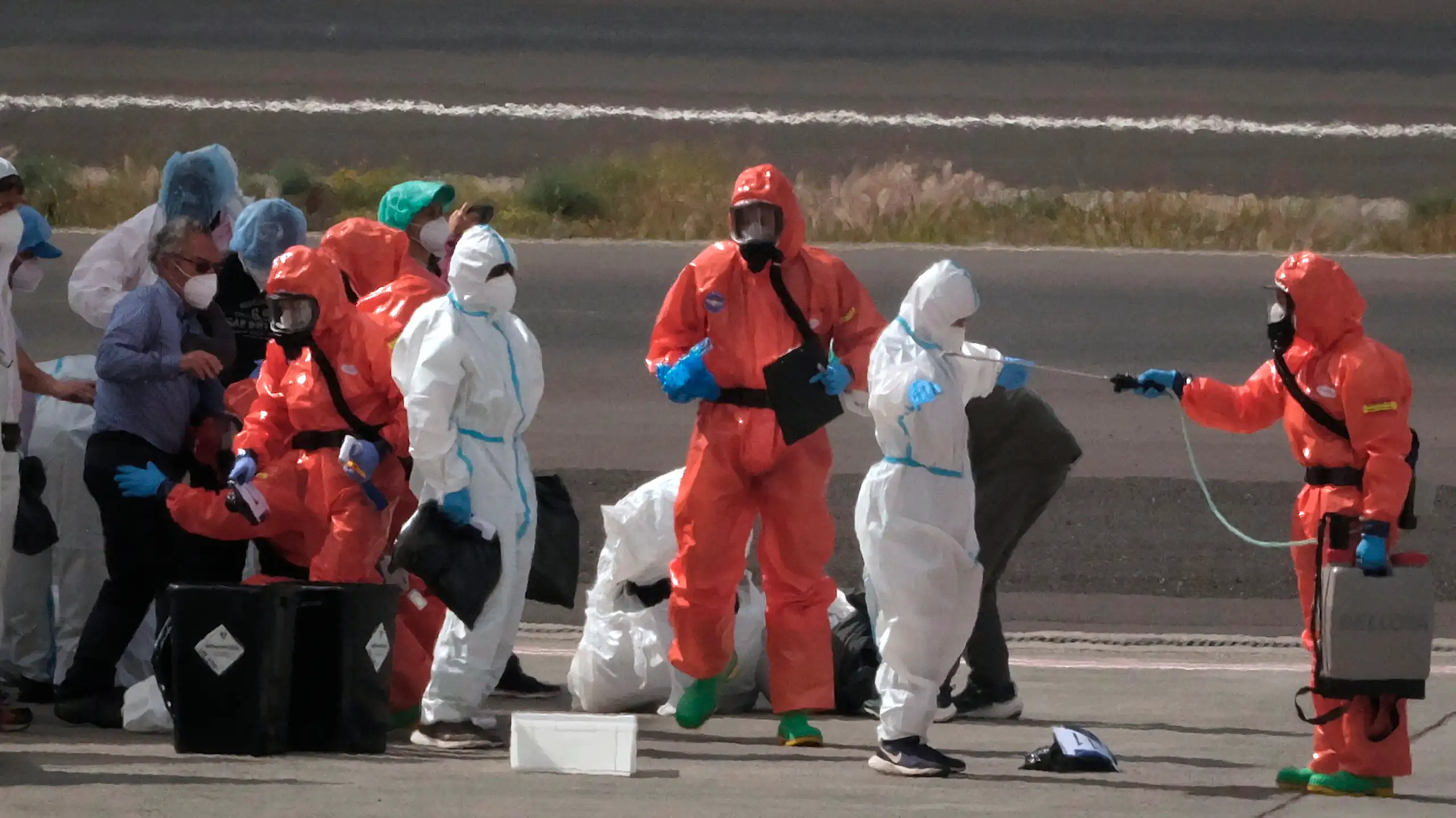 People on a tarmac wearing protective suite, some orange and some white. A person in orange is spraying a person in white while another person in white waits beside an orange-suited person with a clipboard.