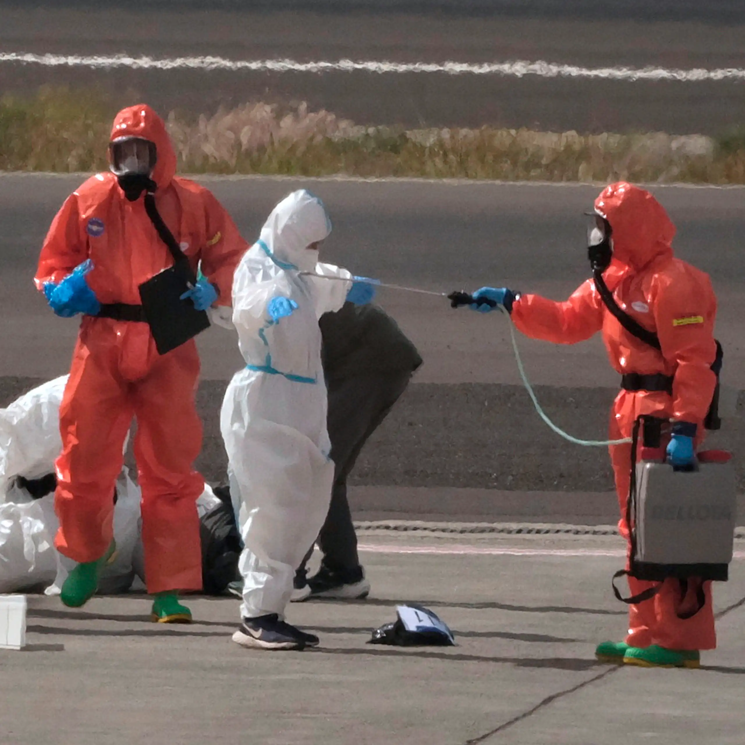 People on a tarmac wearing protective suite, some orange and some white. A person in orange is spraying a person in white while another person in white waits beside an orange-suited person with a clipboard.