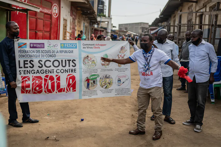Men in face masks hold up an information banner about Ebola while another speaks