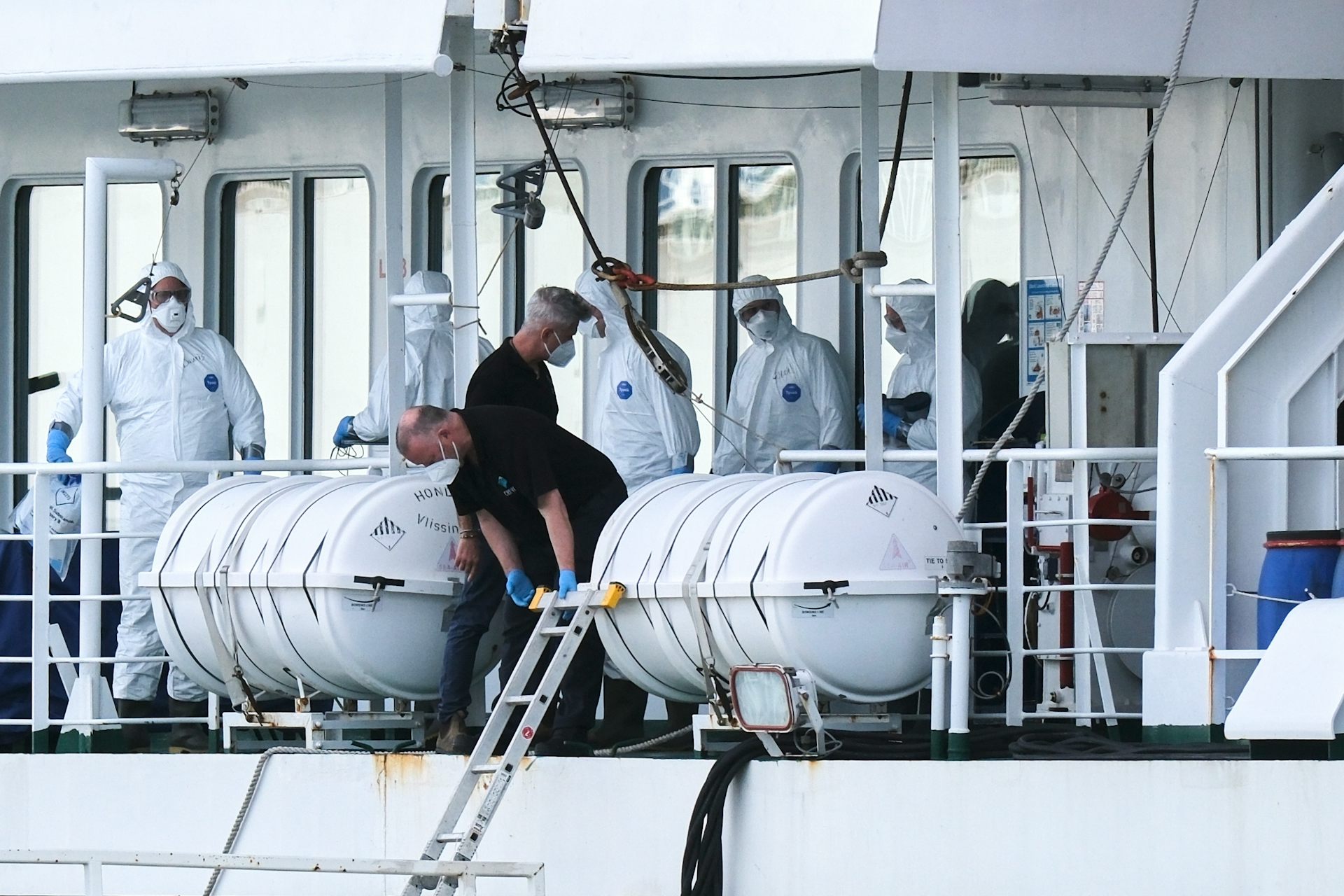 Personas con trajes blancos contra materiales peligrosos en la cubierta de un barco con dos personas con camisas oscuras y máscaras faciales