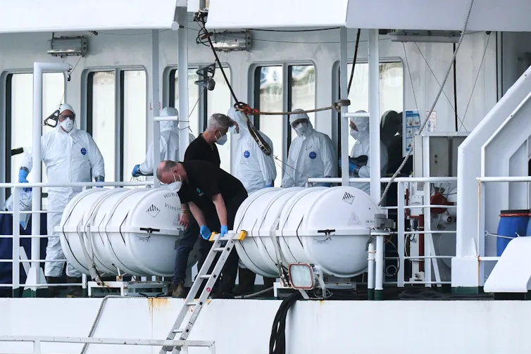 Aviones, trenes y pandemias: lecciones de la COVID-19 sobre los riesgos de viaje que plantean el hantavirus y el ébola 2 Personas con trajes blancos contra materiales peligrosos en la cubierta de un barco con dos personas con camisas oscuras y máscaras faciales