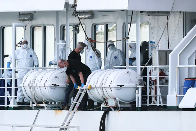 People in white protective suits on the deck of a boat with two people wearing dark shirts and face masks