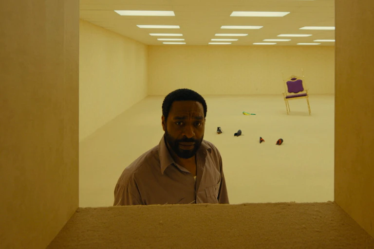 A man in a bland, tan coloured hallway looks towards the camera through a square window.