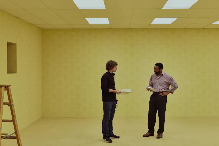 A young director speaks to an actor on a blank, beige-coloured set.
