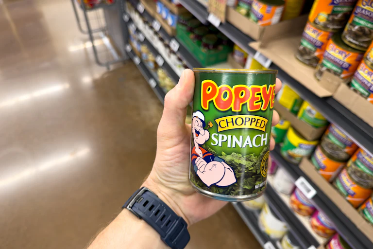 a hand holds a tin of spinach featuring the cartoon character Popeye