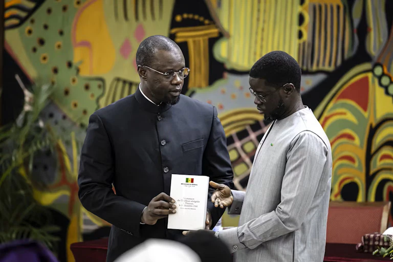 Two African men shake hands, one dressed in black and one in grey.