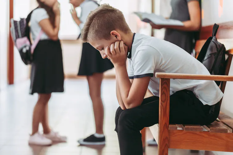young boy sitting alone on a bench in a school with his head in his hands.