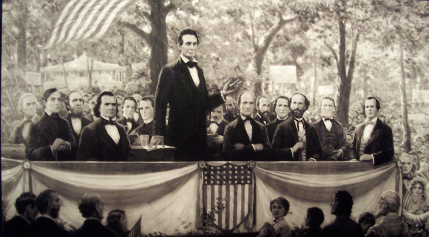 A black and white illustration of around a dozen men in suits, including a standing Abraham Lincoln, on a platform amid a crowd outdoors.