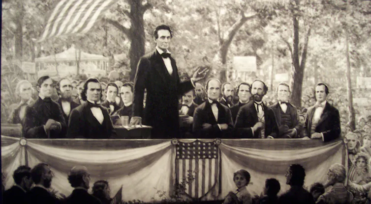A black and white illustration of around a dozen men in suits, including a standing Abraham Lincoln, on a platform amid a crowd outdoors.