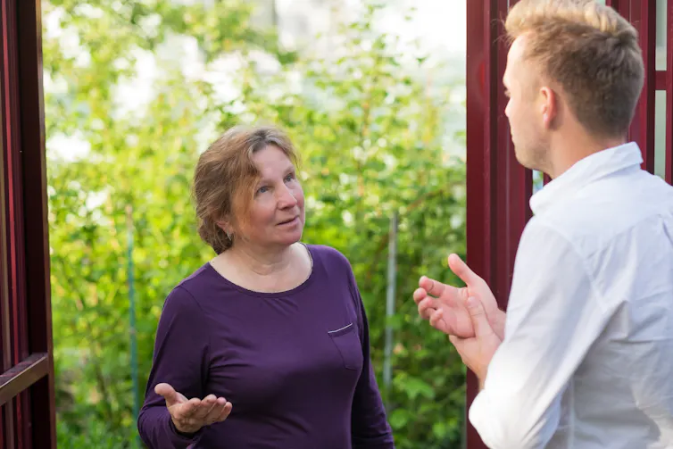 A middle-aged woman in a purple shirt stands in a garden speaking with a tall, younger man in a white collared shirt.