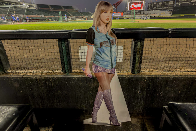 A cardboard cutout of a young blonde woman wearing a teal baseball jersey in the dugout at a baseball stadium.