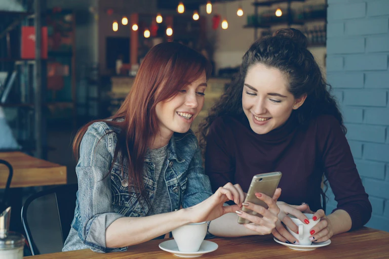 Two young women with long hair sit in a cafe smiling at something on their phone.