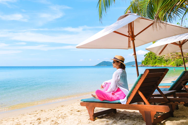 Woman wearing light summer clothes and a hat, sits on a beach on sun lounger under a parasol and looks out to the sea