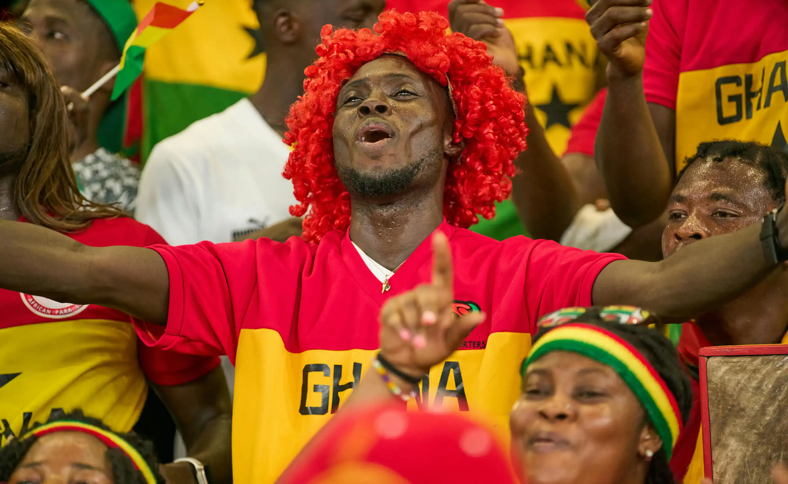 A joyful man in a red wig holds his arms out in a football stand, surrounded by others in brightly coloured outfits.