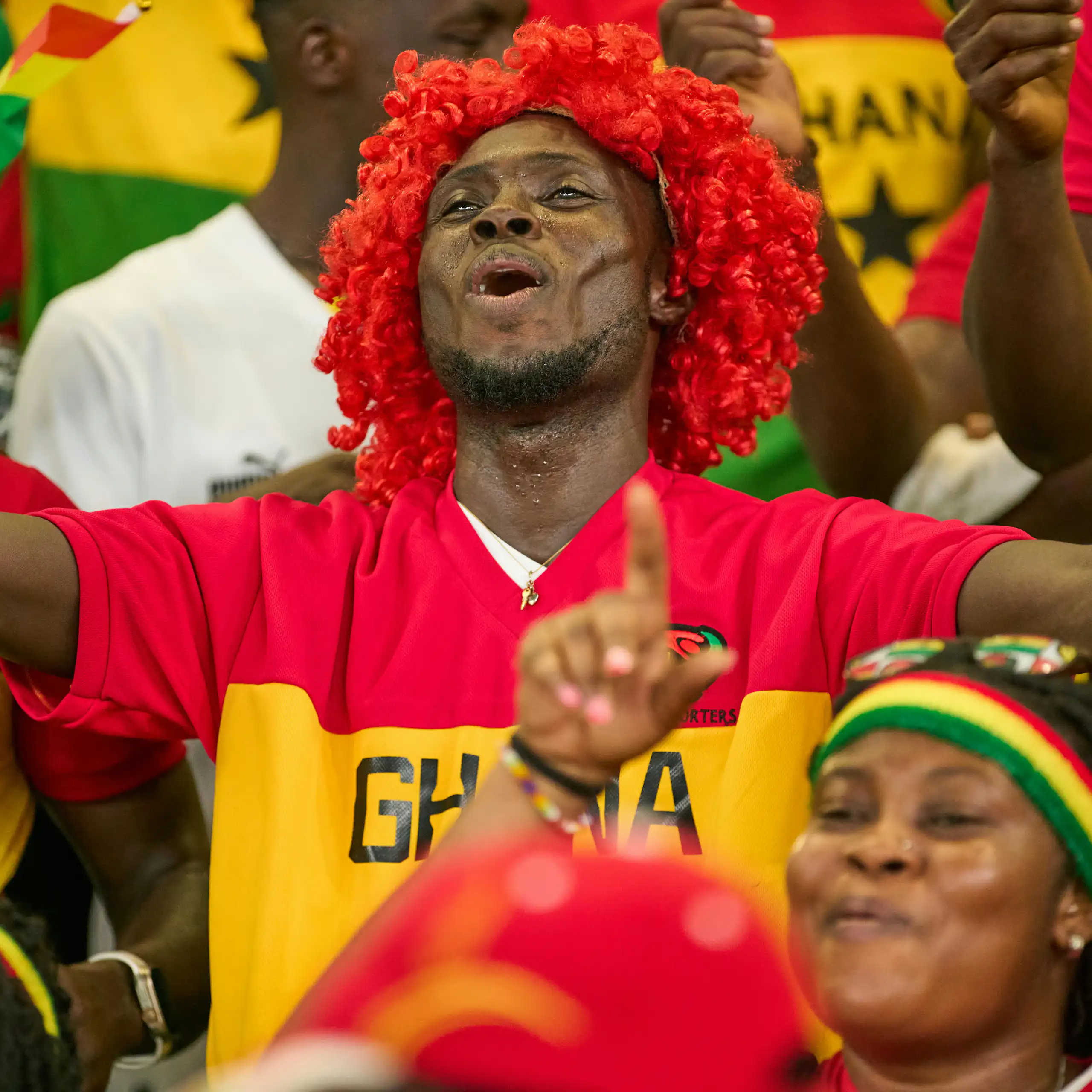 A joyful man in a red wig holds his arms out in a football stand, surrounded by others in brightly coloured outfits.