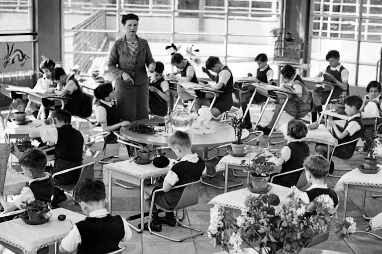 Photo prise en classe : les bureaux des élèves sont disposés en cercle, l'institutrice se tient au centre.