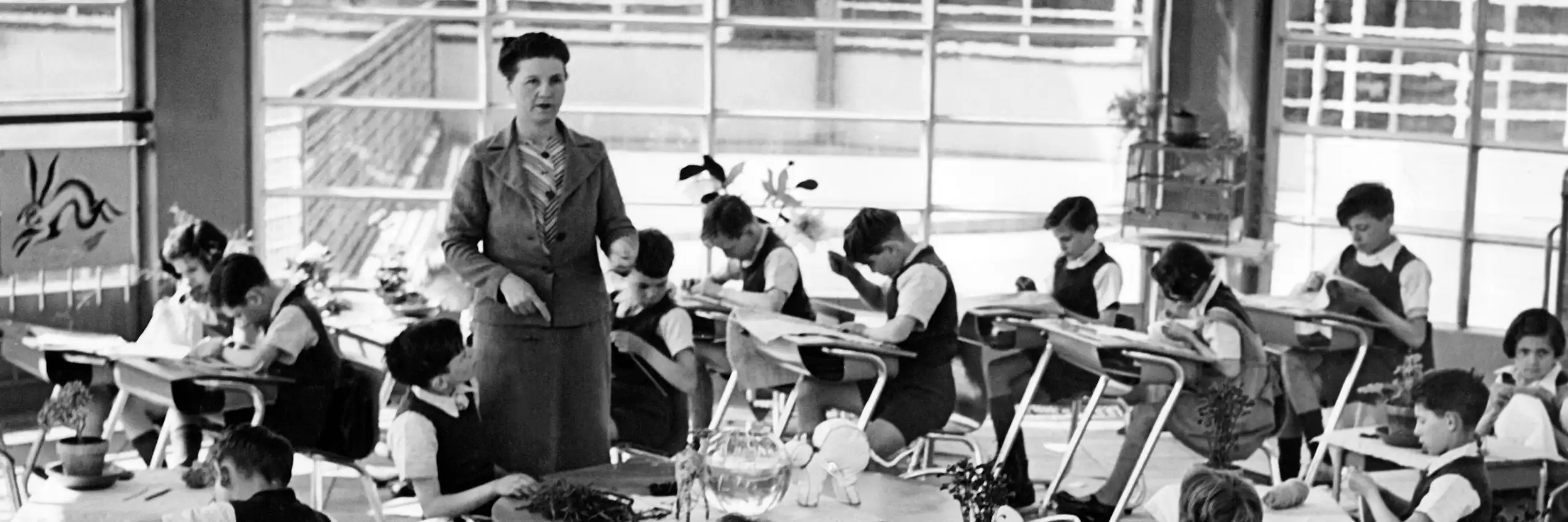 Photo prise en classe : les bureaux des élèves sont disposés en cercle, l'institutrice se tient au centre.
