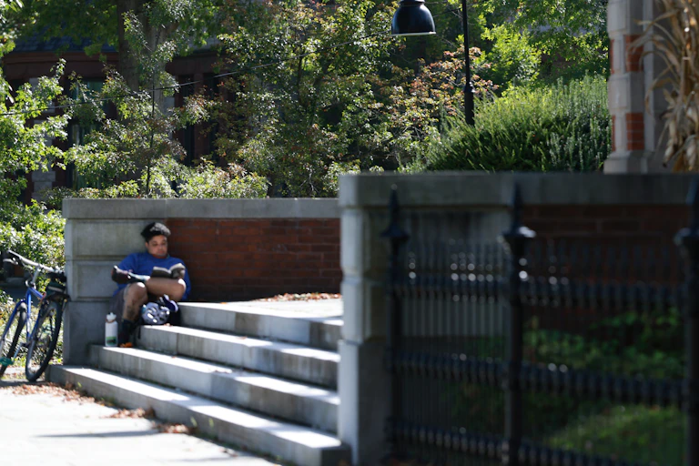 A person sits and reads a book on stone steps, with leafy trees behind them.