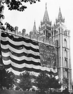 A black and white photo of a tall building with sharp spires, and a striped flag with stars.