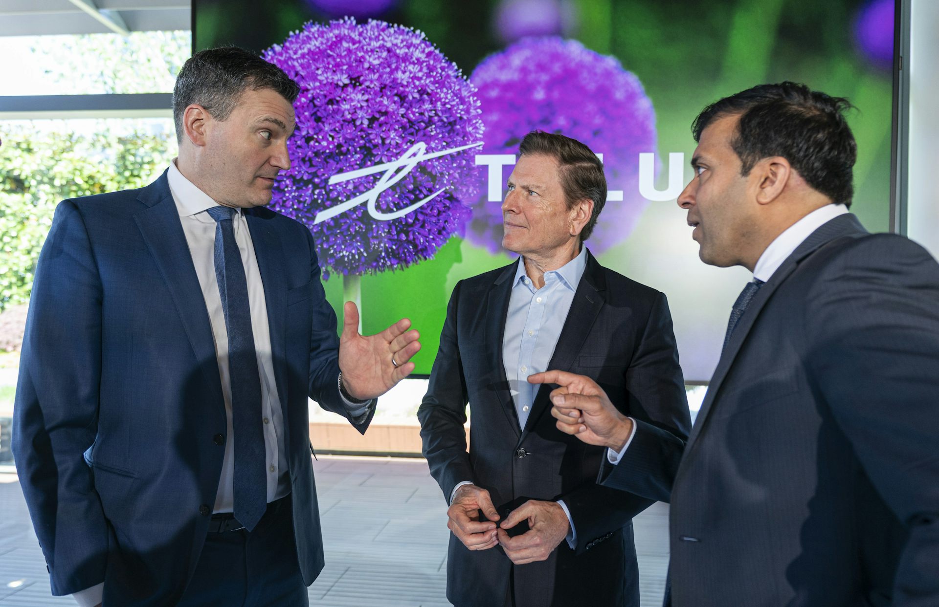 Three men in business suits speak animatedly, with a Telus logo in the background