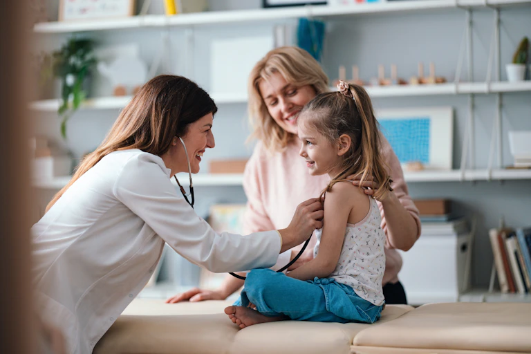 A friendly pediatrician performs a health examination for a happy young girl, with her mother in the background.