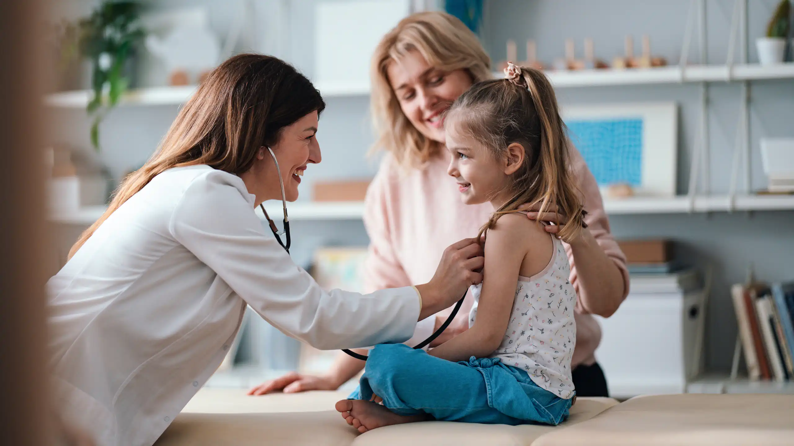 A friendly pediatrician performs a health examination for a happy young girl, with her mother in the background.