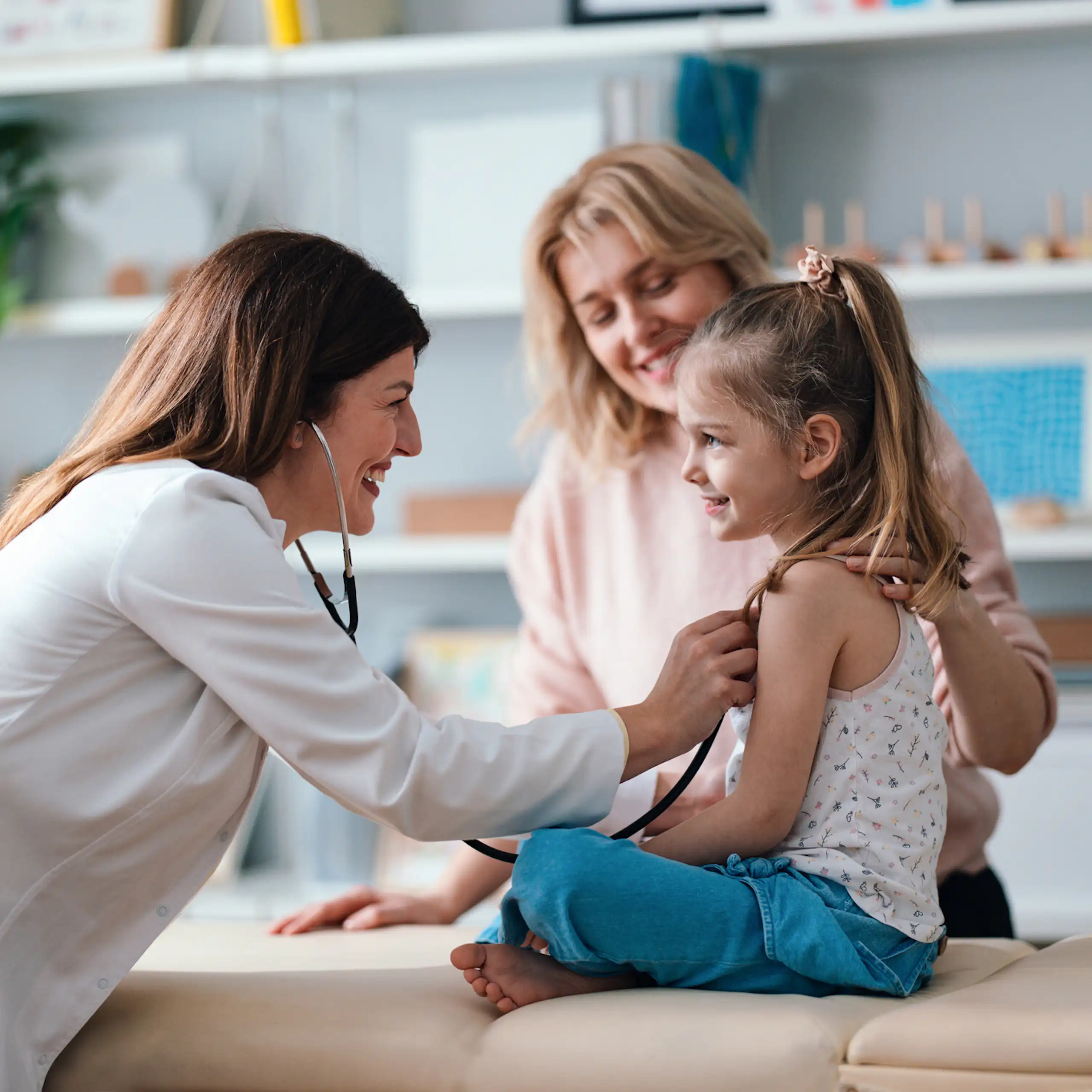 A friendly pediatrician performs a health examination for a happy young girl, with her mother in the background.