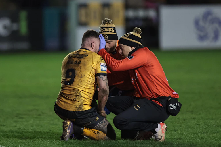 Two men crouch down to examine another on a rugby pitch.