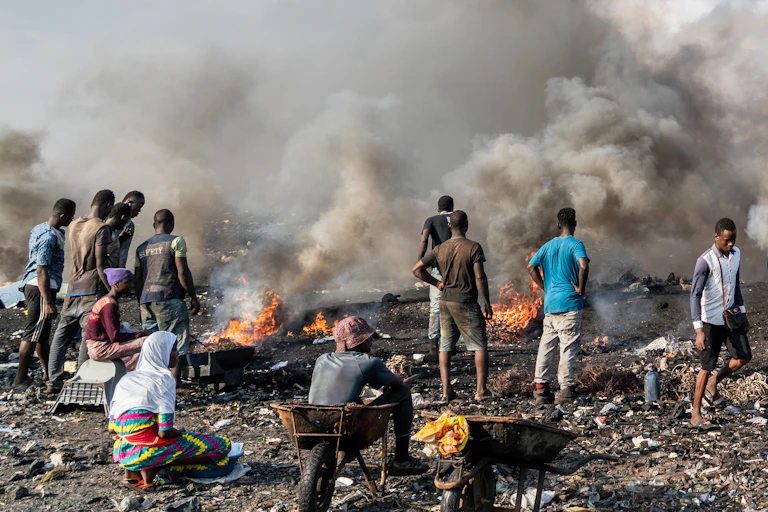 An open air garbage site with fires on it and clouds of smoke; about 35 people are sorting through discarded electronic goods