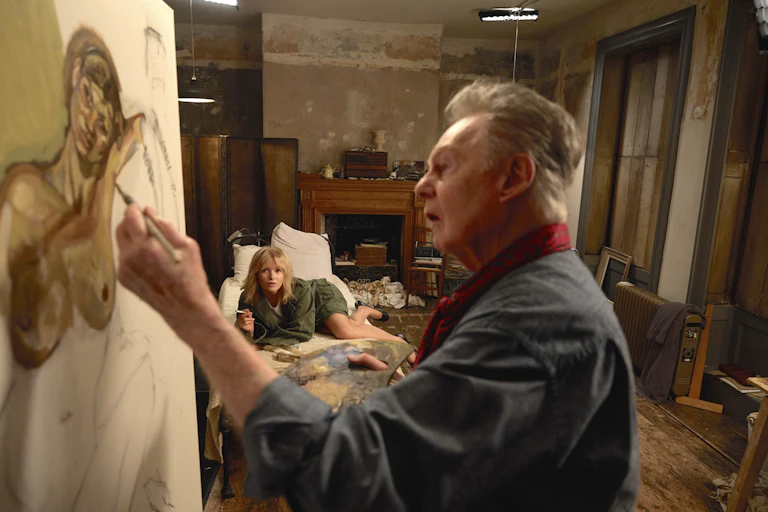 An older man at an easel painting a young woman lying on a bed smoking a cigarette.