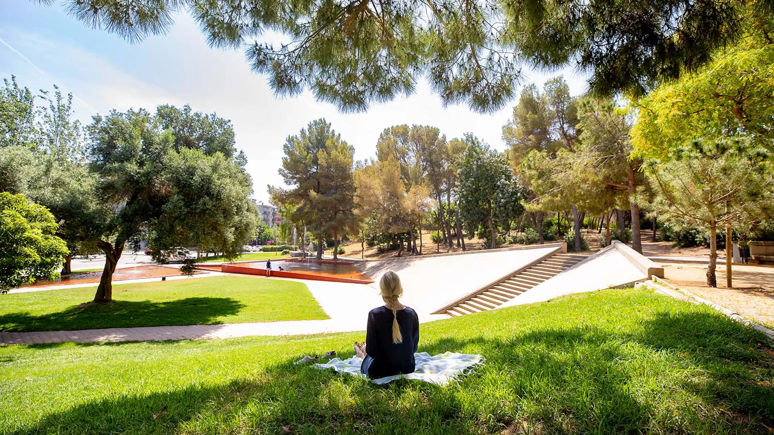 Une femme est assise dos à l'herbe dans un parc où se trouvent des pins et une fontaine en arrière-plan