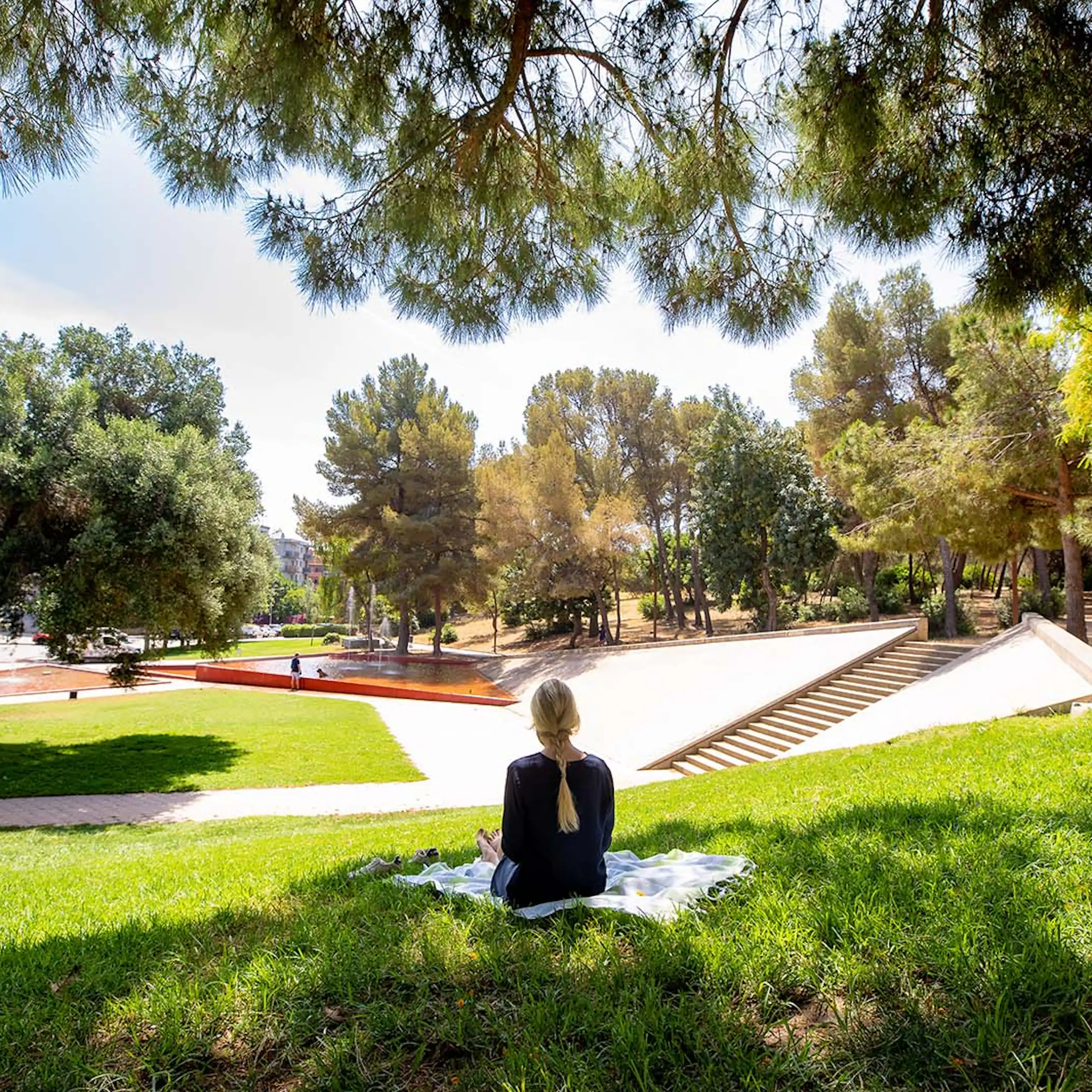 Une femme est assise dos à l'herbe dans un parc où se trouvent des pins et une fontaine en arrière-plan