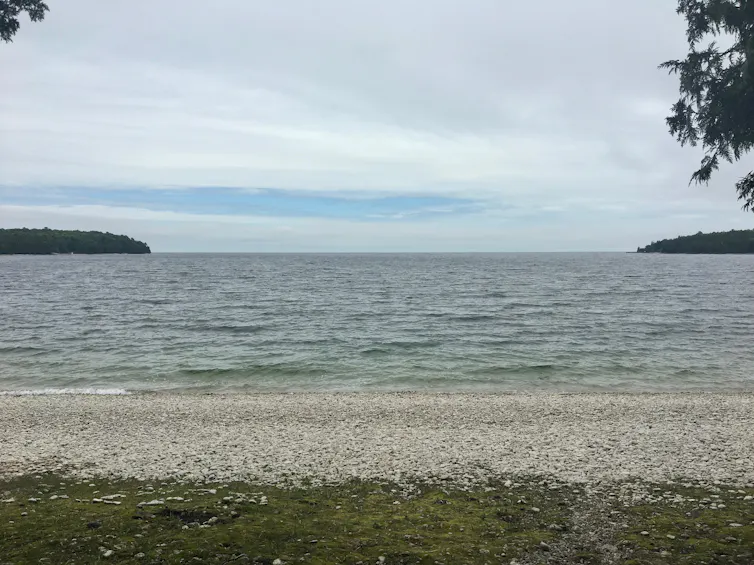 A view of a rocky beach looking out at a lake with tree-covered land in the distance.