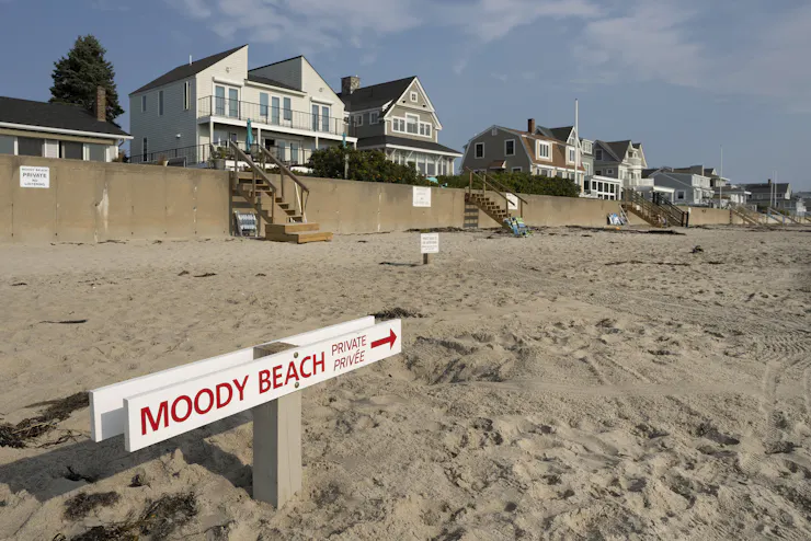 A beach with large homes behind a sea wall also features a sign saying 'Moody Beach - private.'