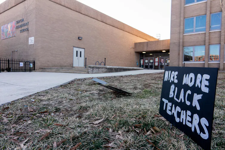 A black sign says 'Hire more Black teachers' on grass outside a red brick building.
