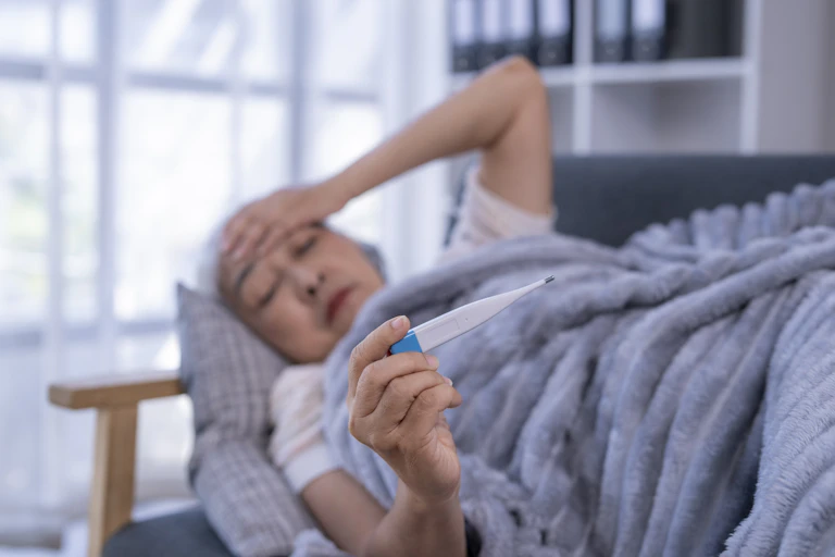 Elderly woman lying under blanket on couch clutching forehead with one hand, holding thermometer in other.
