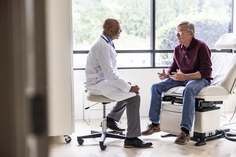 An older male doctor and older male patient talk in a medical exam room.