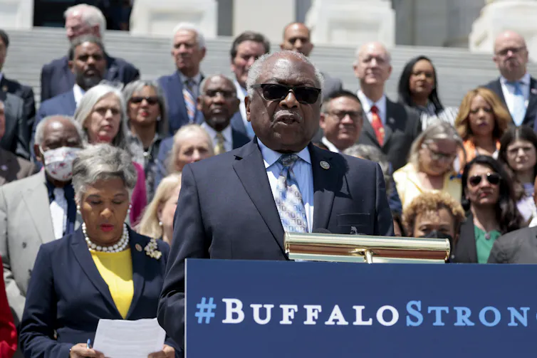 A Black man wearing sunglasses speaks outdoors in front of a lecturn.