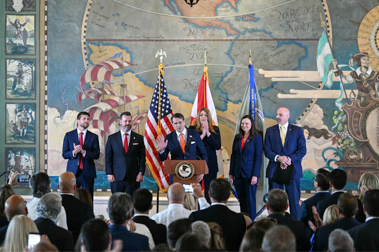 A man in a suit and tie speaks at a lectern as five people stand behind him.