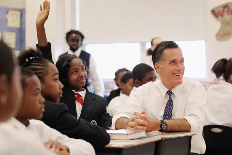 Man in white dress shirt and tie sits at school desk around middle school students in uniforms