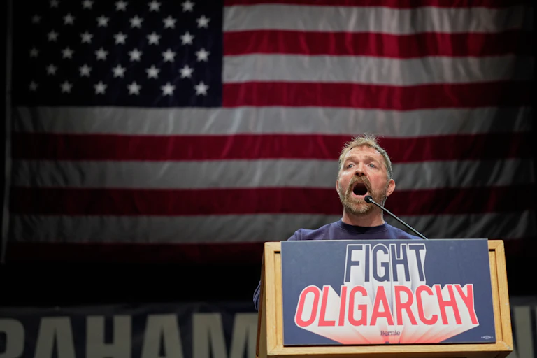 A man at a lectern that has the sign 'FIGHT OLIGARCHY,' in front of a US flag, speaking.